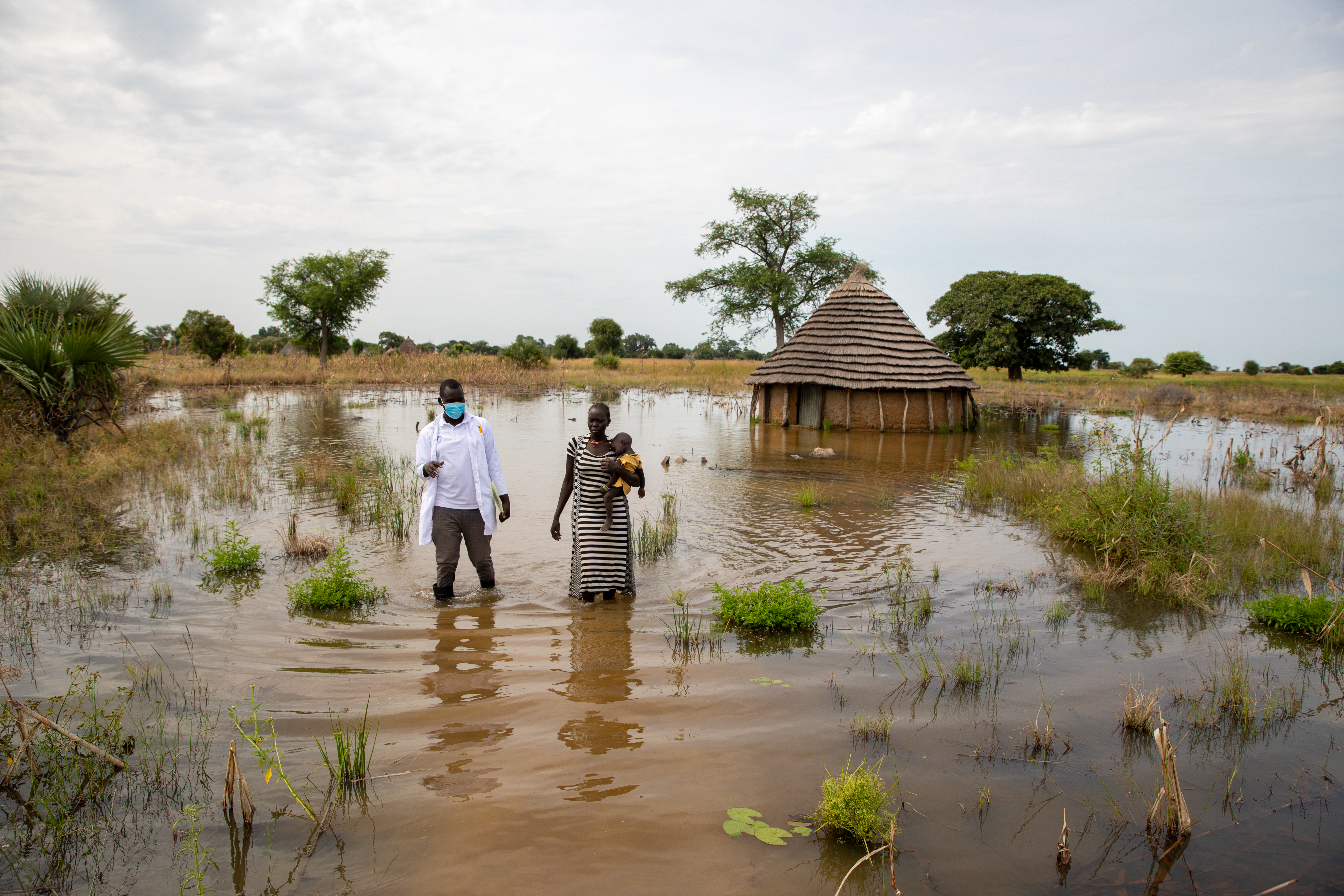 James from IRC and Abuk, holding a sleeping Nyirou walk in the floods surrounding Abuk's tukul, in South Sudan